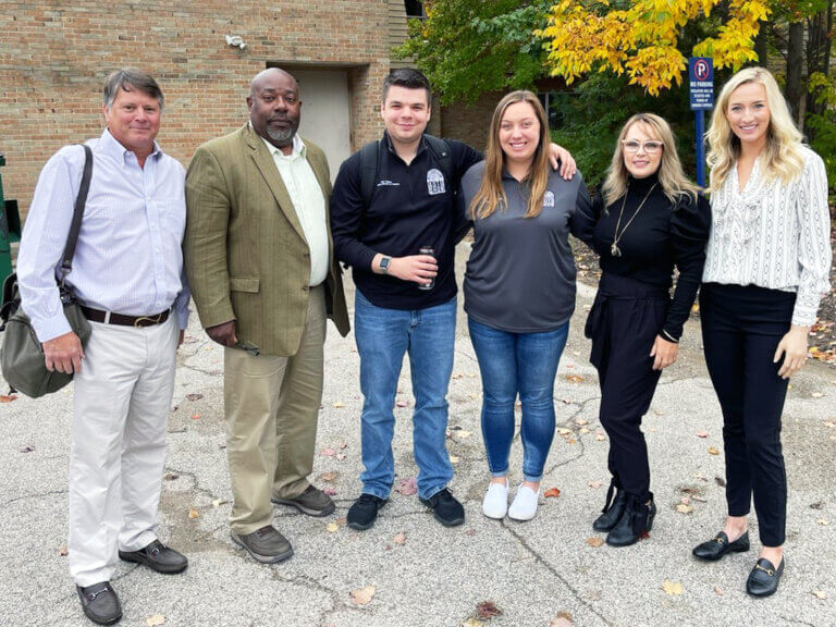 A group of six individuals standing side by side outdoors, dressed in a mix of casual and semi-formal attire.