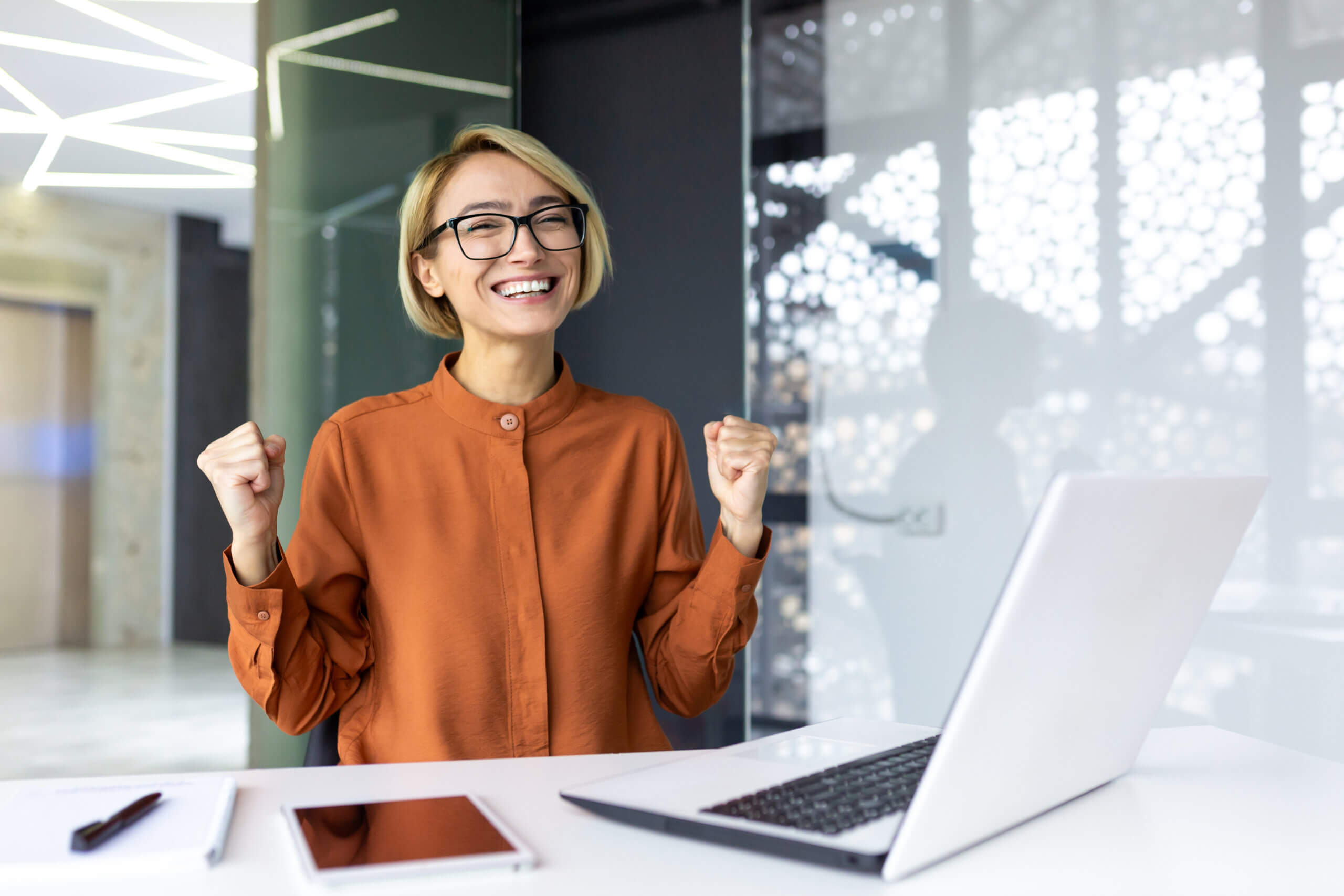 Young joyful business woman boss smiling and looking at camera, employee celebrating victory and successful achievement results looking at camera holding hands up triumph gesture