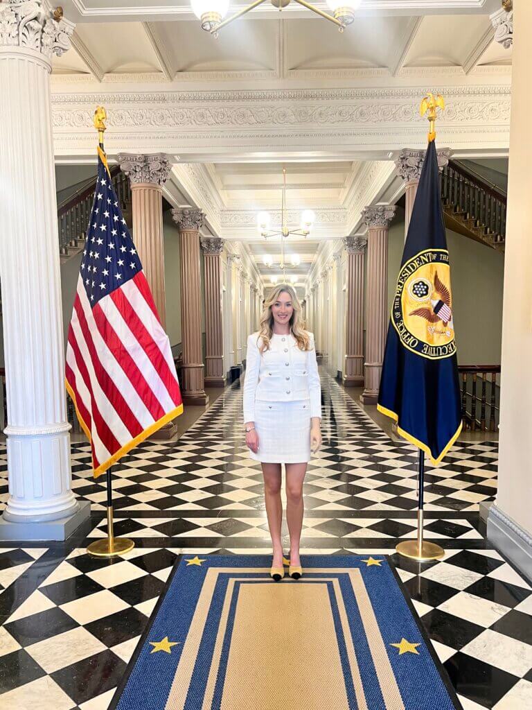 An individual stands in the center of a hallway with a checkered black and white floor. On either side are the United States flag and another flag with an emblem. The hallway features ornate columns and decorative ceilings.