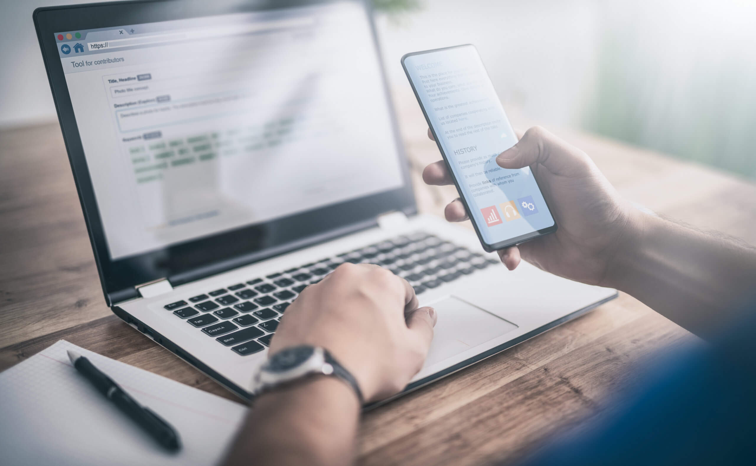 Man at desk crafts an email on his desktop as he looks at his mobile phone