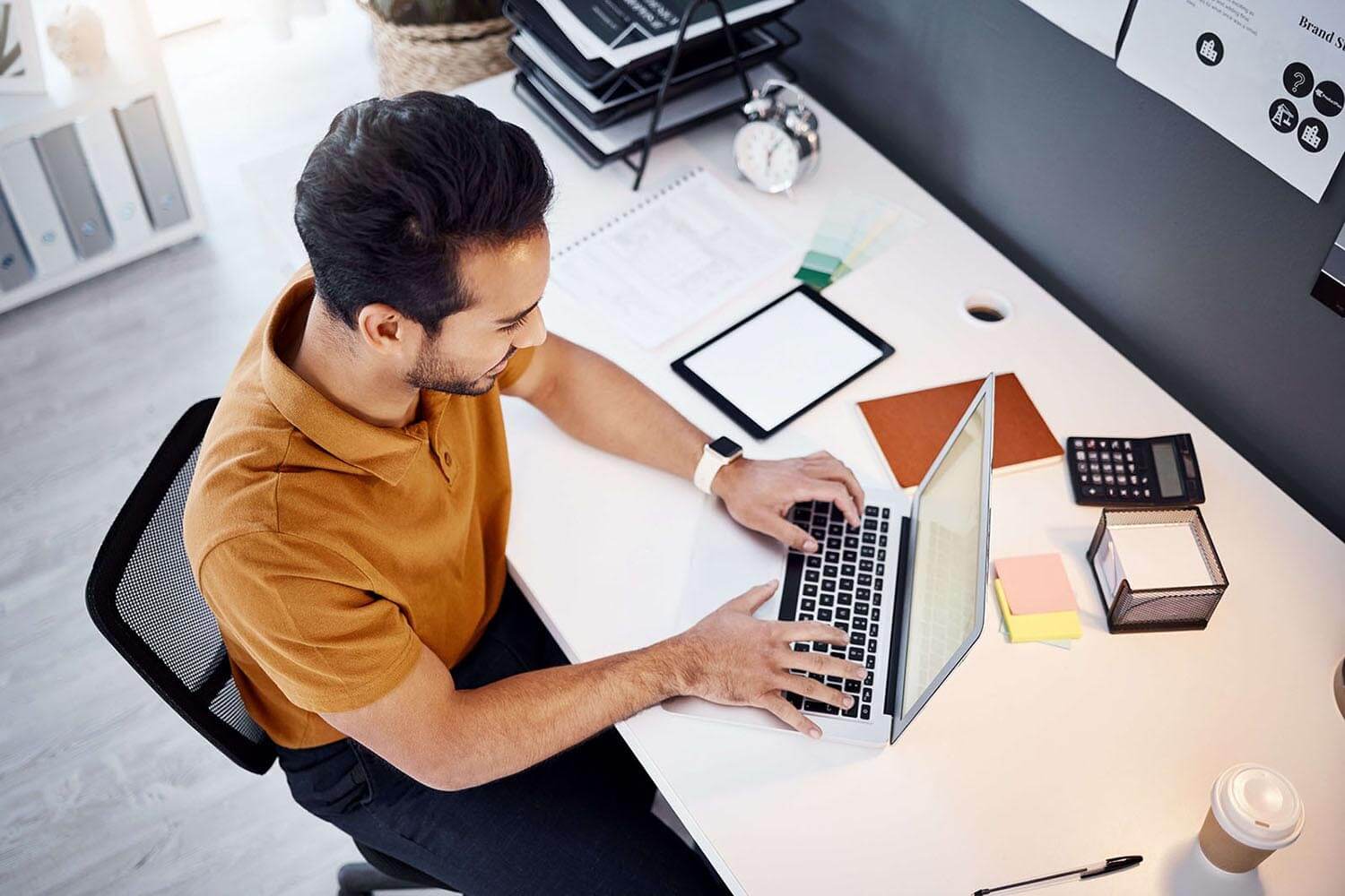 Man at a desk working on a laptop with office supplies around.