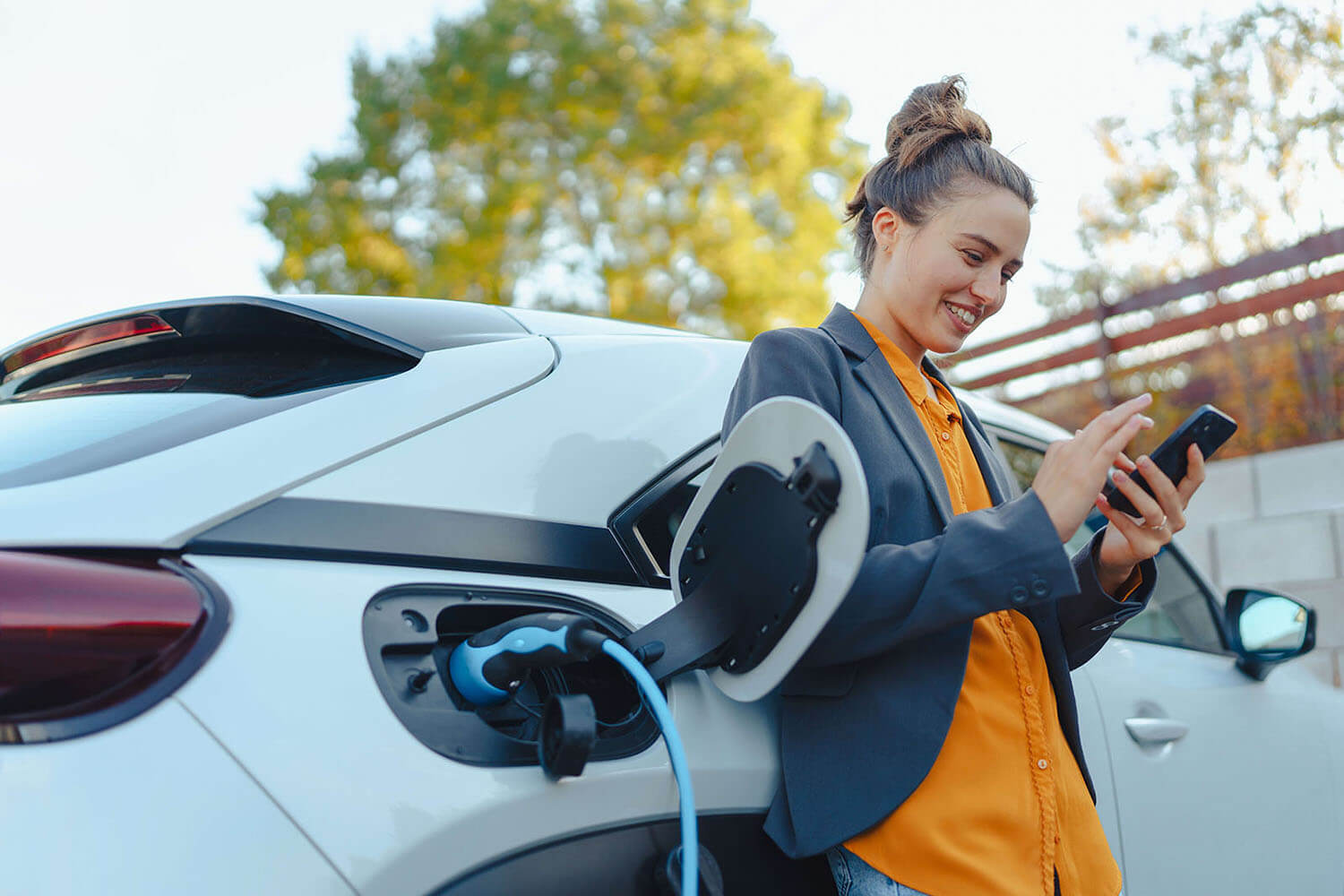 Young smiling woman leans on a charging EV as she checks her phone.