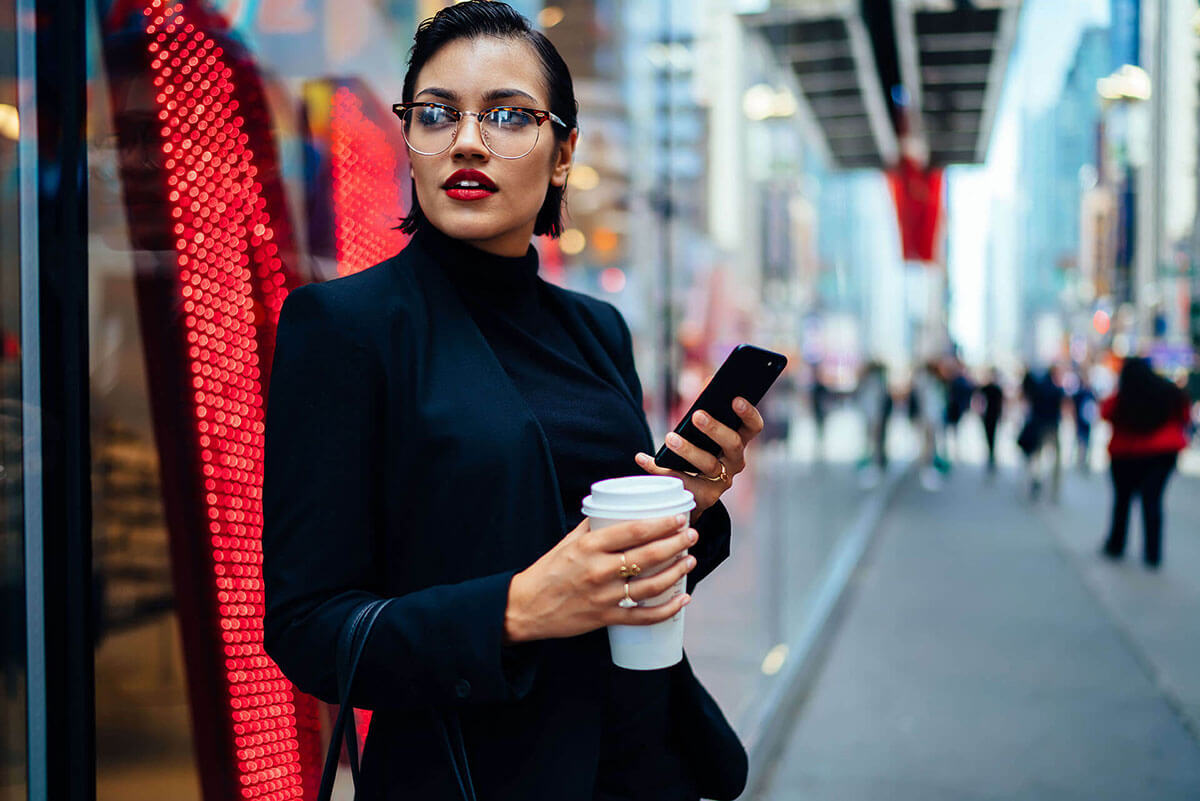 Stylish female employee on the go walks down a sidewalk interacting with her phone.