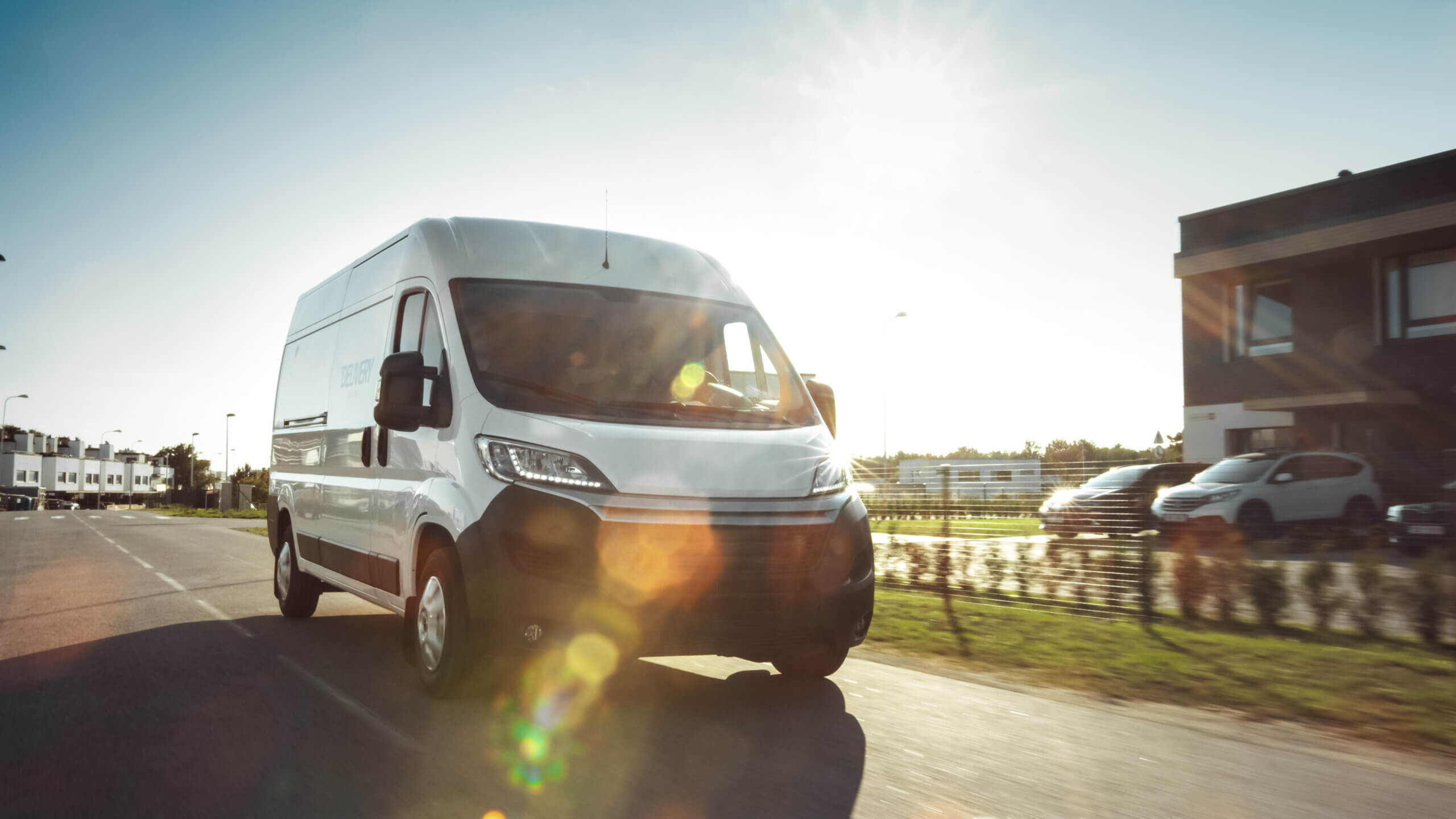 A white commercial vehicle driving on an urban road with clear skies and sunlight causing lens flare