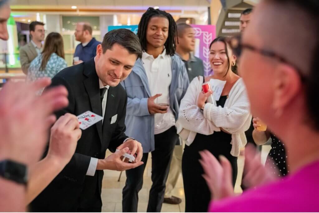 A magician in a black suit performs a card trick for a small group, smiling as the audience watches closely. One woman laughs, holding a cup, at an indoor social event