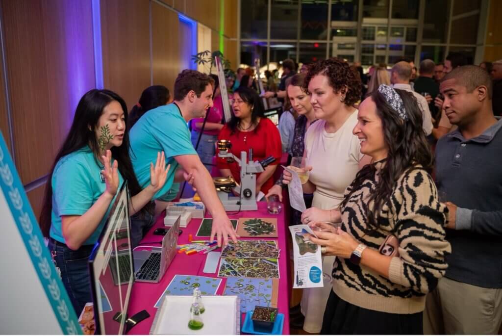 Two presenters in turquoise shirts engage with a group of attendees at an exhibit booth, showcasing scientific materials, including microscopes and plant samples.