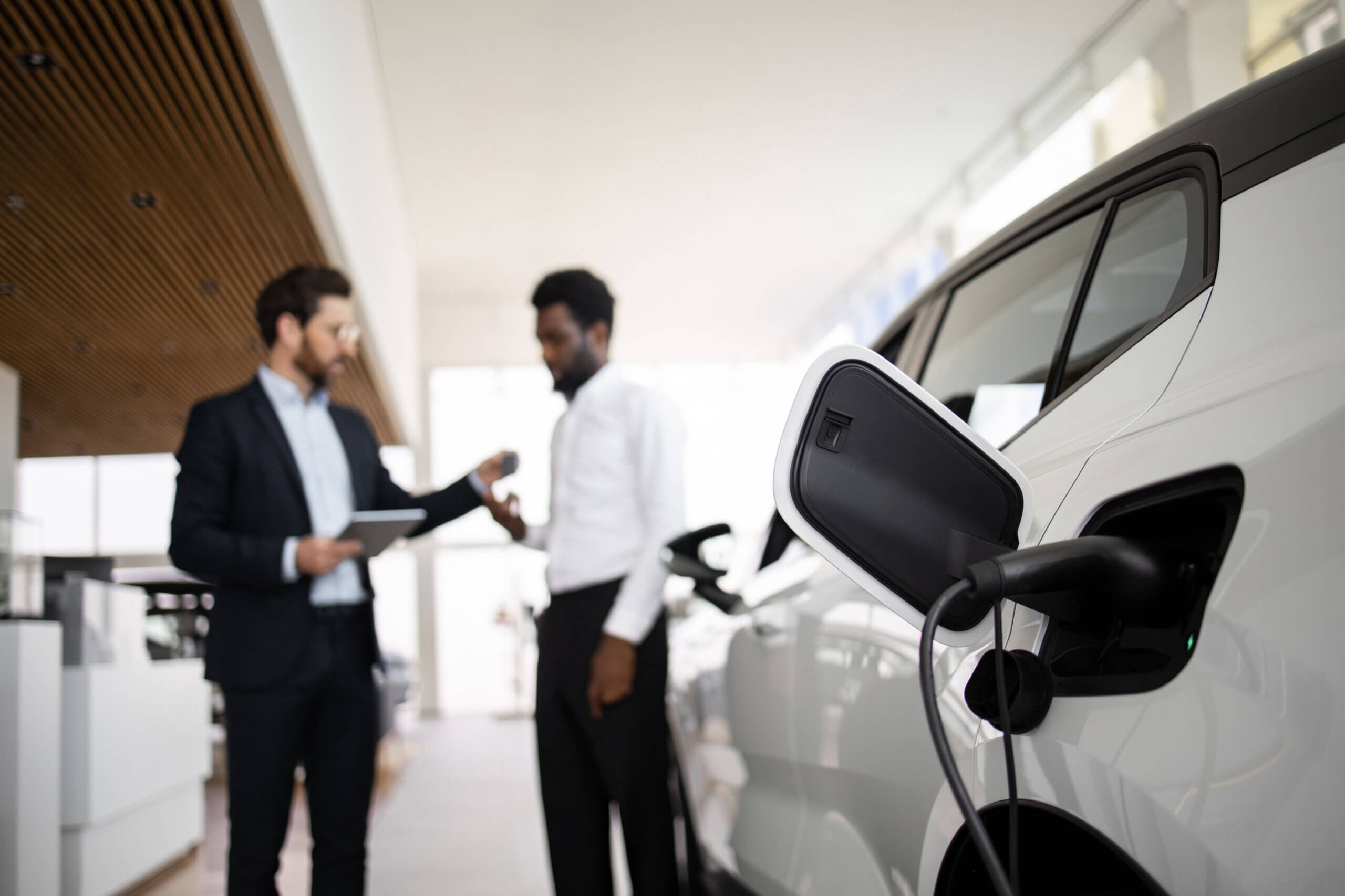 Two men in a modern showroom discuss an electric vehicle charging at a station. One man is holding a tablet, while the other is gesturing toward the car. The focus is on the car's charging port and cable.