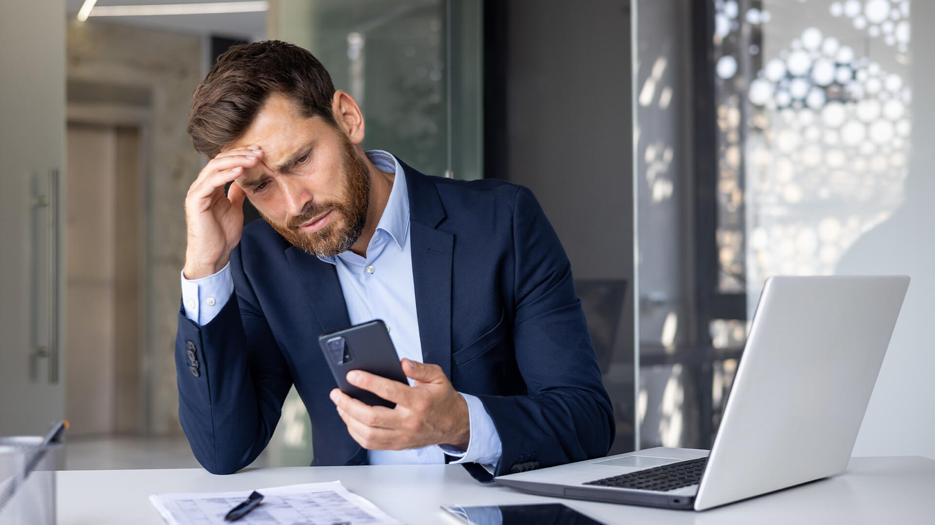 A person in a business suit sits at a desk with a laptop, holding a smartphone and resting their head on one hand.