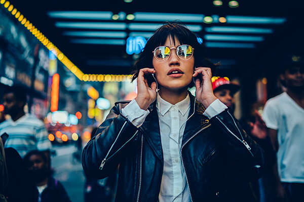 Half length portrait of trendy female millennial in electronic spectacles looking at camera during time for listening audio book and walk in metropolitan downtown, generation z in earphones