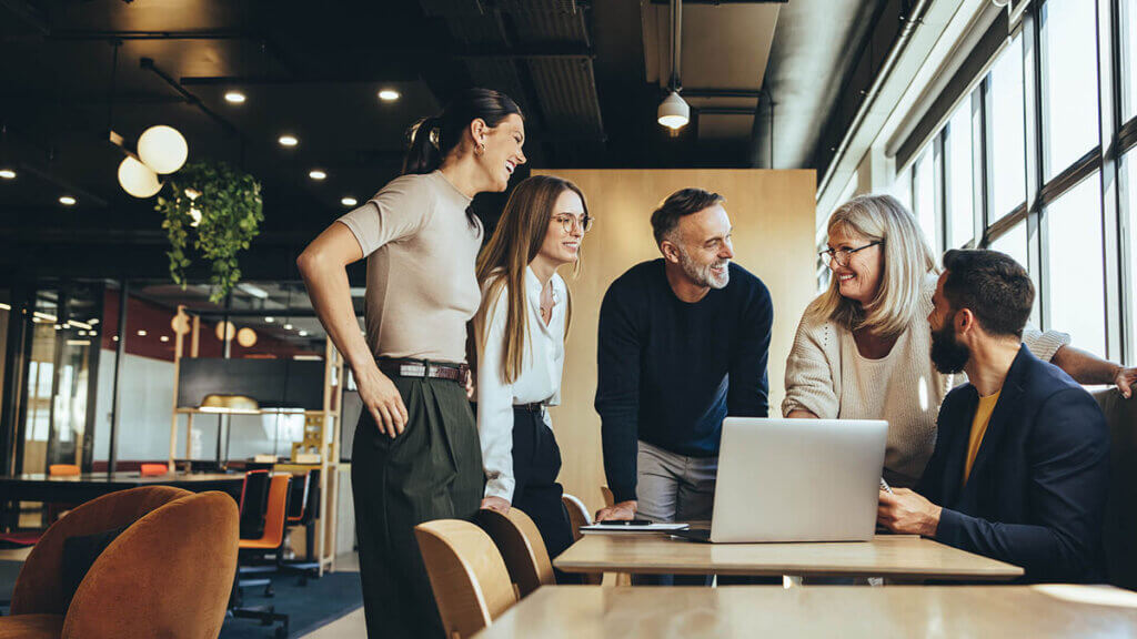 A group of five people gathered around a table in an office setting, engaged in a discussion and smiling. A laptop is open on the table.