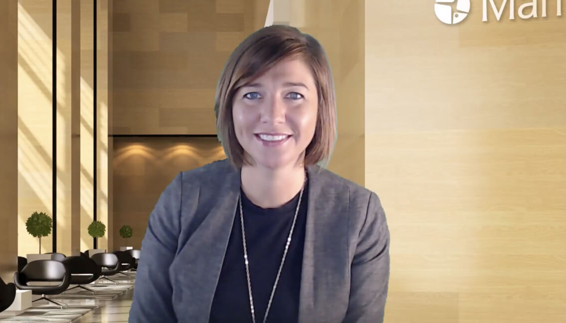 A person with short hair wearing a grey blazer and black shirt is smiling at the camera. The background shows a modern office lobby with wooden walls, black chairs, small green plants, and large windows letting in natural light. The logo "Maritz" is partially visible in the top right corner.
