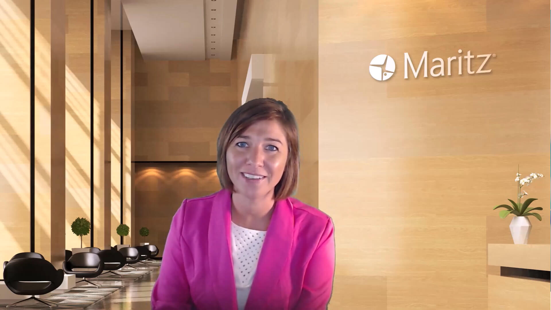 Person wearing a bright pink blazer standing in a modern office lobby with beige walls, black chairs, potted plants, and the Maritz logo displayed on the wall.