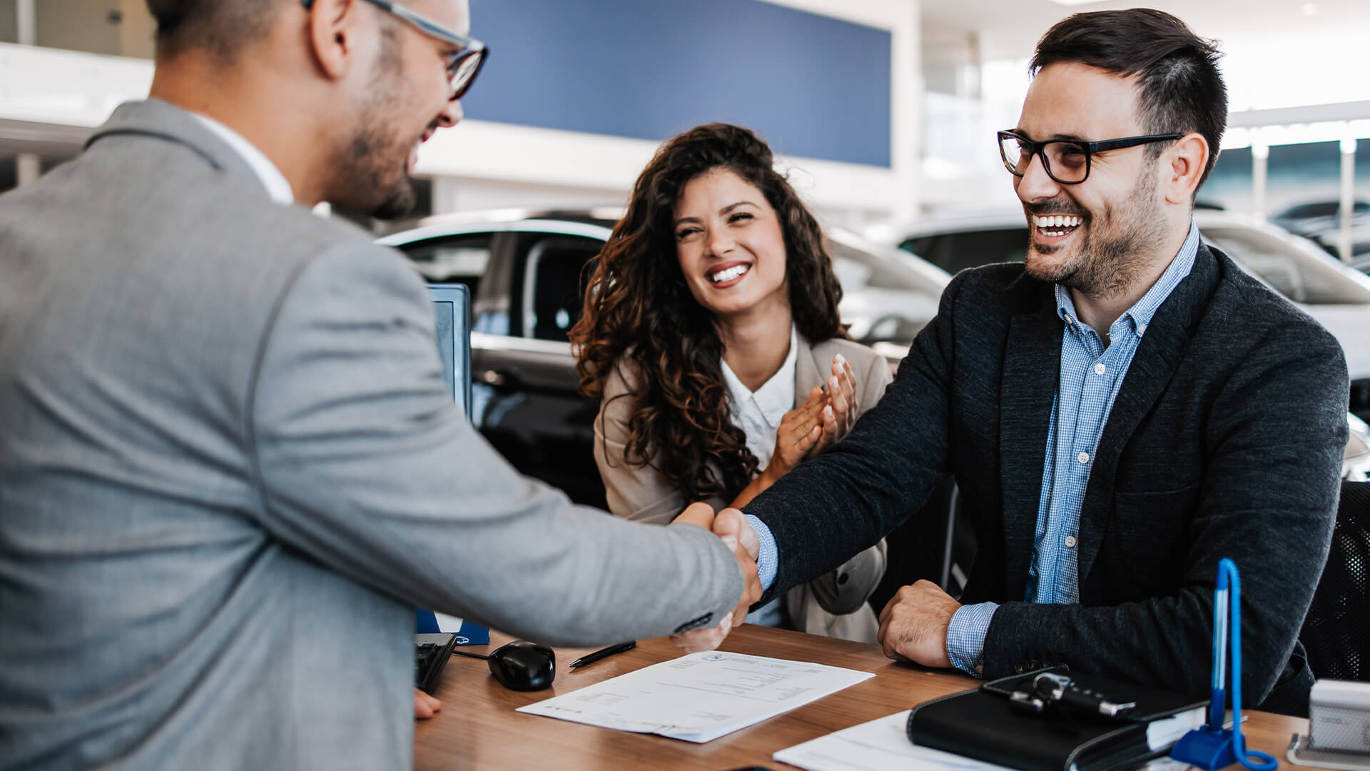 Two individuals shaking hands across a desk with paperwork and car keys, inside a modern car dealership.