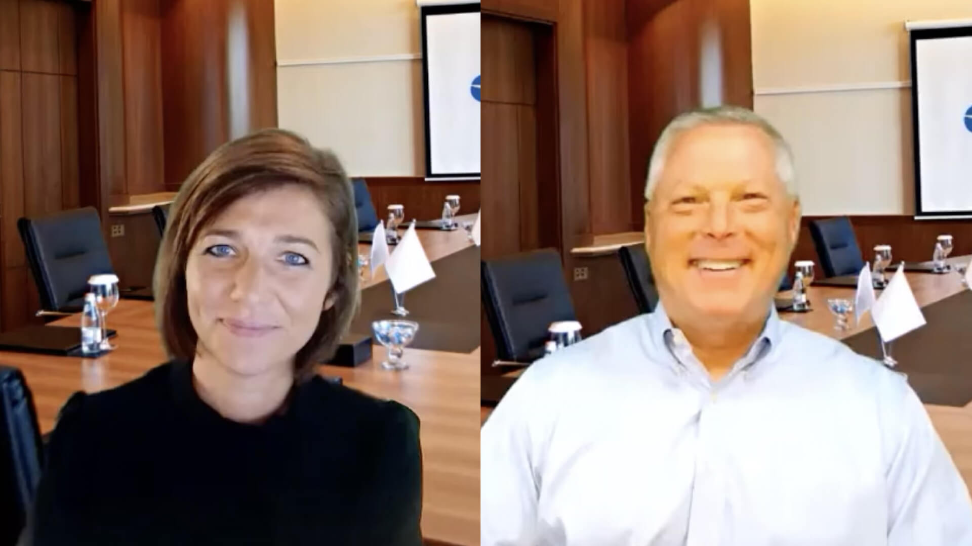 Two people shown side by side in a conference room setting with a boardroom table, chairs, and presentation screen in the background.